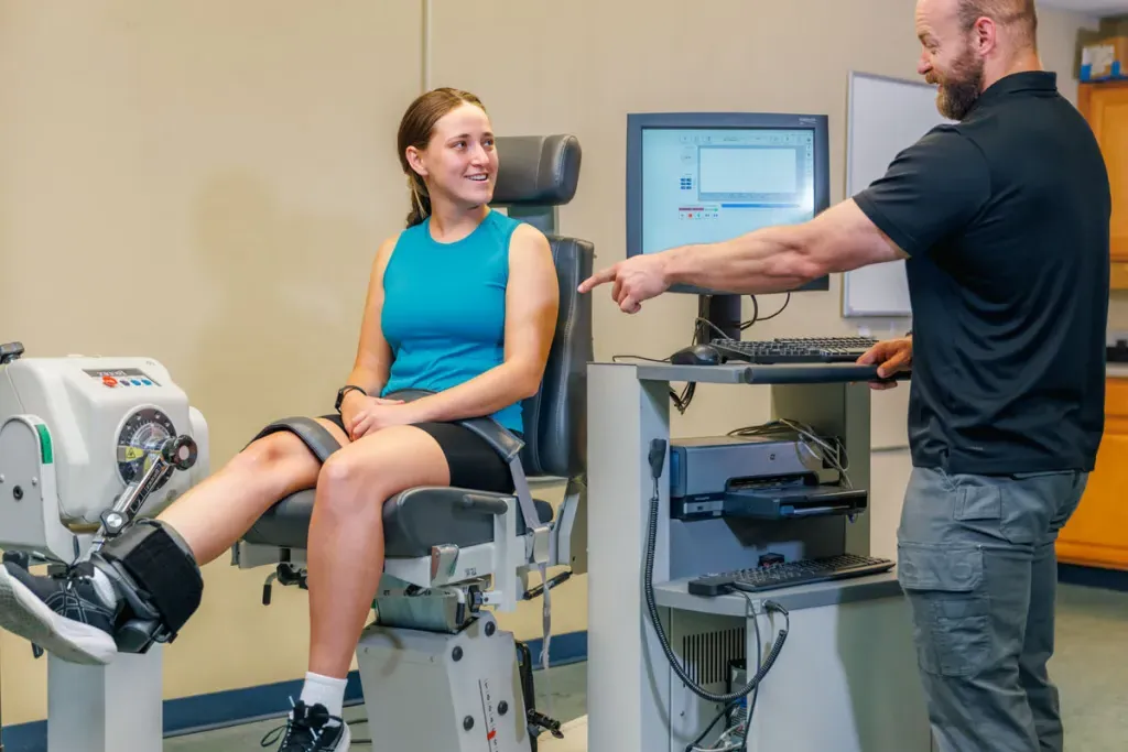 A student sits in a machine that measures muscle strength as their professor shows them the results on a monitor