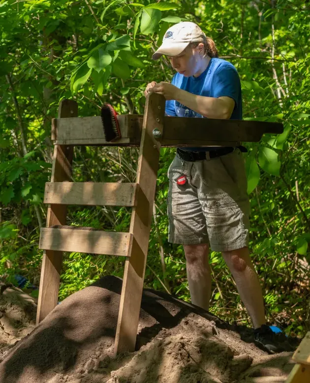 A student stands at a sifting table above a dig site